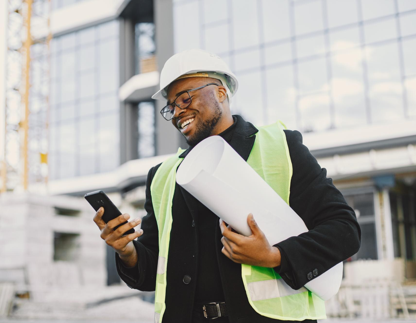 Construction professional with hard hat and safety vest looking at smartphone while holding blueprints